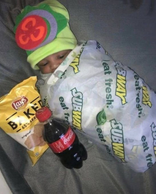 A baby wearing a colorful beanie is bundled up in a Subway wrapper with chips and a bottle of Coca-Cola beside them.