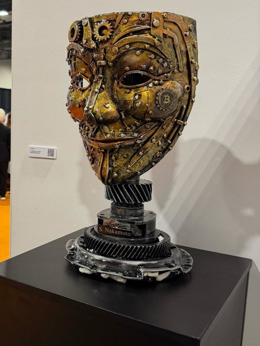 A steampunk-style sculpted head made of metal gears and mechanical parts sits on a black pedestal labeled 'S. Nakamoto,' displayed in what appears to be an art exhibition.