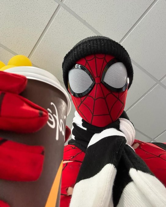A low-angle selfie of someone wearing a Spider-Man mask, black beanie, and striped scarf, holding a McDonald's McCafe cup with red-gloved hands.