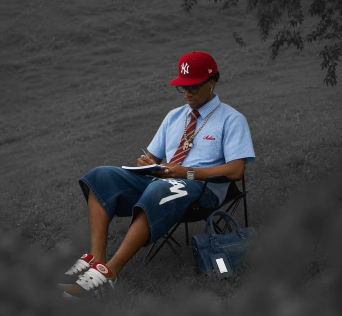 A person in a red NY cap, sunglasses, striped shirt, tie, long denim shorts, and sneakers sits writing in a notebook outdoors.