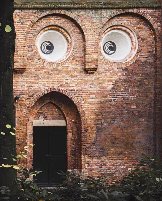 An old brick wall features a dark arched doorway and two large circular windows designed to look like cartoon eyes, with concentric circles drawn on them.