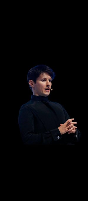 A photograph of a man with dark hair wearing a black turtleneck, standing against a dark background with his hands clasped together, appearing to be speaking or presenting.