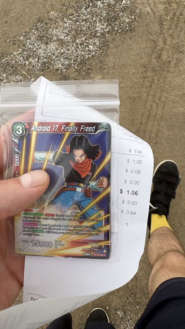 A hand holding a Dragon Ball Super trading card titled 'Android 17, Finally Freed' worth $1.06 according to the receipt, taken at an outdoor retail location.