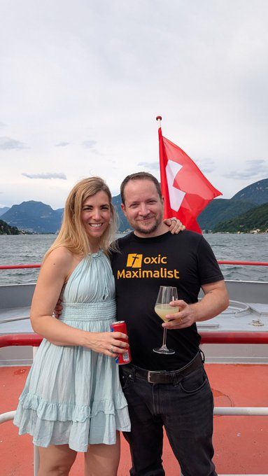 A couple poses on a boat with a Swiss flag behind them, the woman in a light blue dress holding a red can and the man wearing a black "toxic Maximalist" t-shirt holding a wine glass, as they cap off the LuganoPlanB Summer School 2026 on the lake.