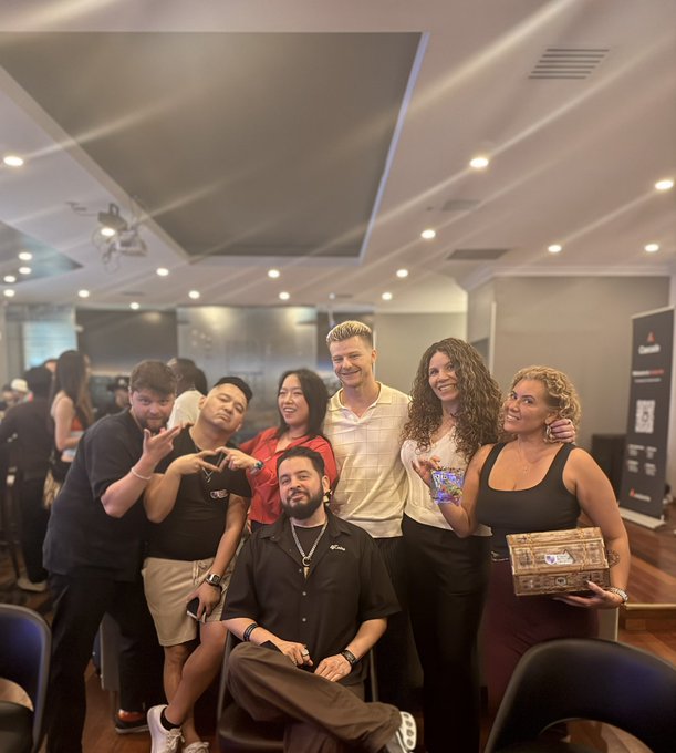 Group photograph of seven smiling people posing together at what appears to be a social or networking event in a modern venue, with one person holding a decorative box.