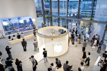 The image shows an indoor event space with people gathered around a large circular display featuring a white structure with a black frame. The room has a high ceiling, allowing for natural light to enter from multiple windows that provide a view of the city outside. There are several people standing and sitting in various positions throughout the room, some closer to the display while others are further away. A few individuals can be seen holding cell phones or taking photos with them. The overall atmosphere appears to be one of interest and engagement as attendees interact with the exhibit and each other.