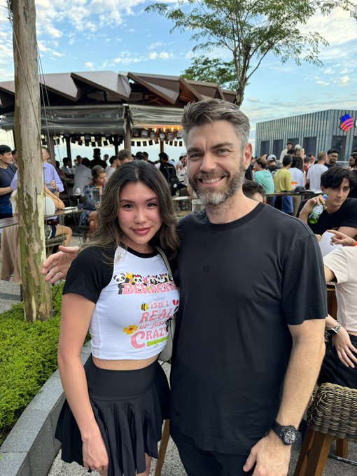 A young woman and a smiling man with a beard pose for a photo together at an outdoor gathering or event.