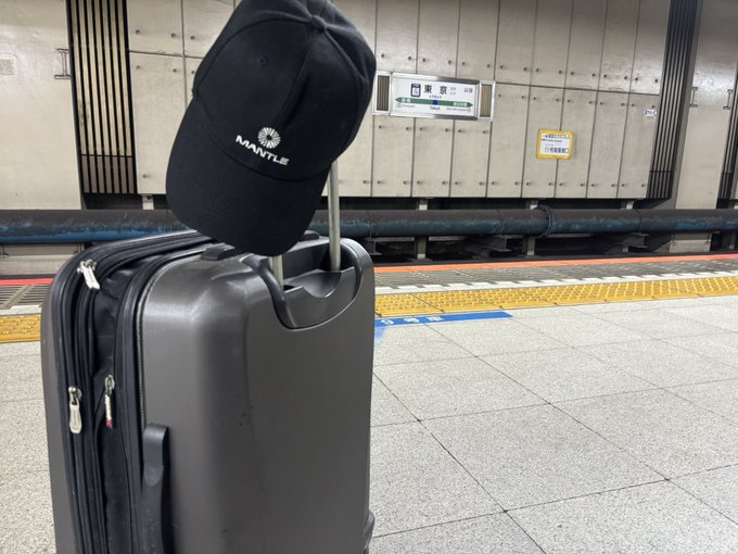 A black Mantle-branded baseball cap hanging from the extended handle of a black rolling suitcase on a subway platform.