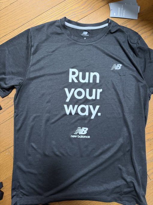Black New Balance t-shirt with 'Run your way.' printed in white text on the front, displayed on a wooden floor with the tag still attached.