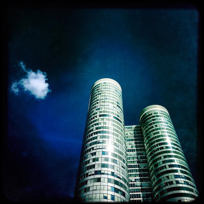 A dramatic low-angle photograph of twin cylindrical glass skyscrapers set against a deep blue sky with a single white cloud, finished with a vintage, vignetted filter.