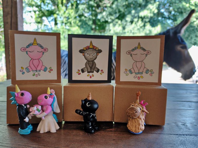 A charming display of three "Unicorn Fart Dust" ($UFD) themed figurines—a wedding couple, a ninja cat, and a llama—arranged on kraft boxes alongside matching illustrated art prints featuring unicorn characters. The caption "Check these out! Community Family $UFD Very Cool @CharleyMakes" ties directly to the collectible merchandise, celebrating the playful, community-driven spirit of the $UFD brand through its whimsical, handmade-style crafts.