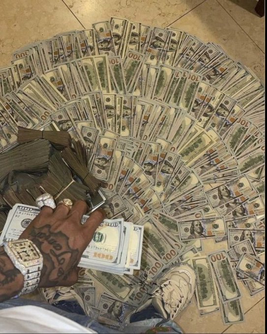 A tattooed hand wearing diamond rings and a luxury watch rests on a large spread of hundred-dollar bills fanned out across the floor, alongside banded stacks of cash and additional bundles.