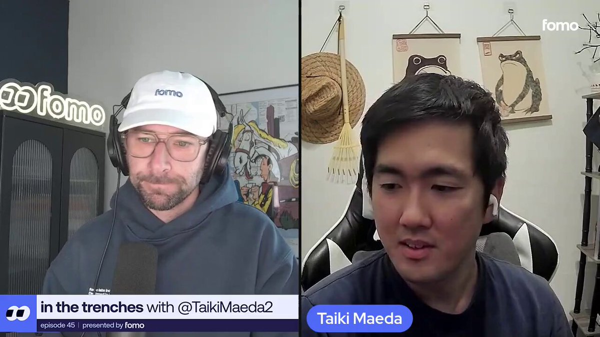 The image shows two men sitting behind microphones during a podcast recording session. The man on the left wears a blue hoodie with a white hat and has his face partially visible through the microphone. The other man, wearing a black shirt, sits across from him in front of a wall adorned with various paintings. They are both focused on the camera, likely discussing the topic at hand or sharing their thoughts about it.