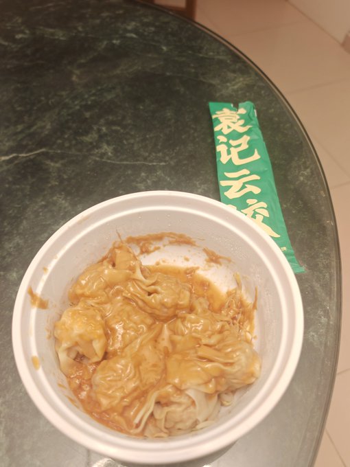A white plastic bowl containing wide rice noodles (cheung fun) coated in a brown sauce sits on a dark marble table next to a green wrapper with yellow Chinese characters.