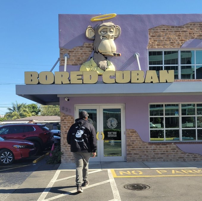A person wearing a "Bored Ape Yacht Club" hoodie approaches the entrance of the "Bored Cuban" establishment, which features a large mural of a Bored Ape NFT character wearing a halo, embodying the proud club membership referenced in the caption.