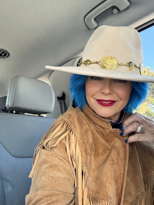 A woman with bright blue hair wearing a cream cowboy hat with a gold band and a tan fringed suede jacket, sitting in a car.