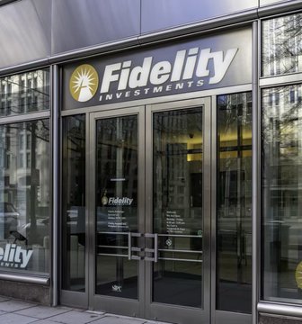 An exterior photograph of a Fidelity Investments office entrance, showing the company's gold sunburst logo and name on glass doors with reflections of surrounding buildings.