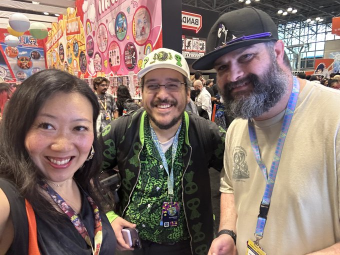 Three people smiling at what appears to be a convention, with a man in the center wearing a Luigi hat and green patterned shirt, flanked by a woman and a bearded man in a black cap.