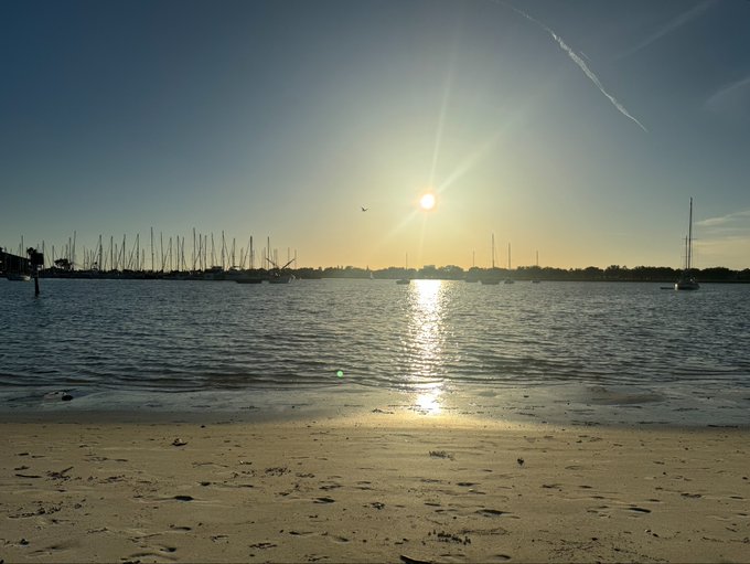 A scenic sunset over a harbor or marina, with the sun reflecting on calm water, silhouetted sailboats and masts along the horizon, and a sandy beach in the foreground.