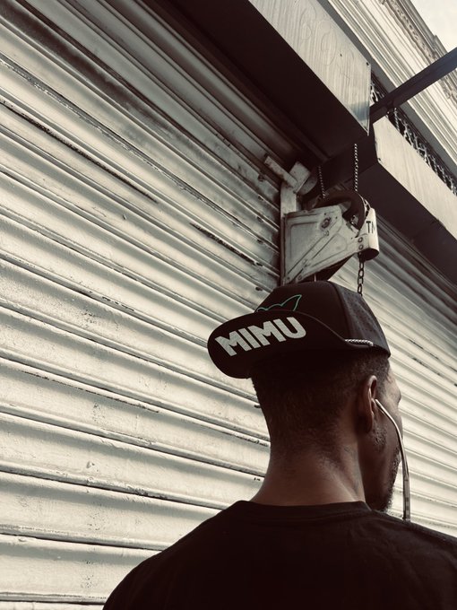 A photograph from behind of a person wearing a black 'MIMU' cap standing near a corrugated metal shutter with a mechanical hoist, wearing earphones.