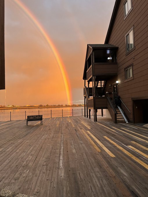 A stunning double rainbow arcs over a wooden deck attached to a brown-sided building, with a sign reading '...' and an exterior staircase visible.