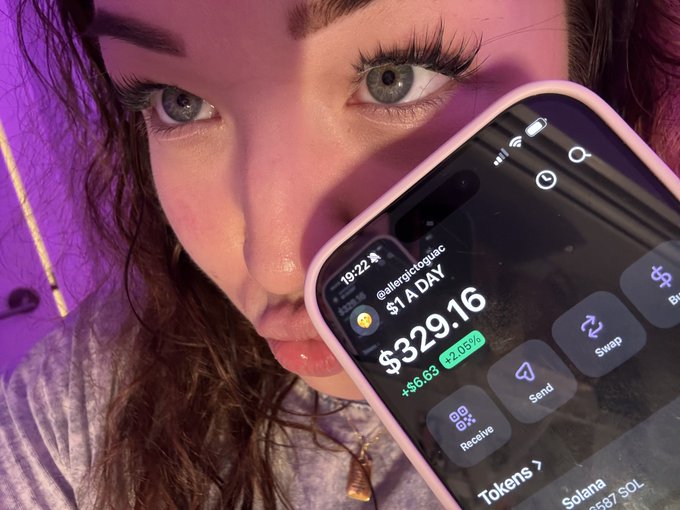 A close-up selfie of a woman showing her eyes and nose, holding a smartphone displaying a cryptocurrency portfolio with the handle '@allergictoquac', a '$1A DAY' contribution, and a total of '$329.16' (up +$6.63 or +2.03%).