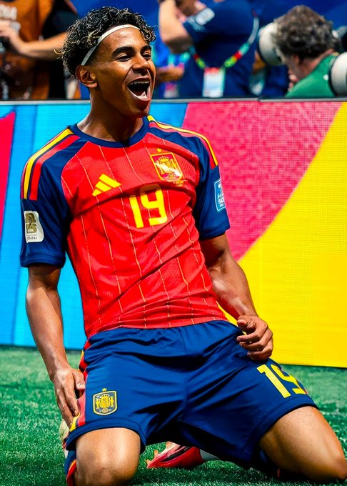 A young soccer player wearing a red and blue Spain jersey with the number 19, kneeling on the grass and shouting in celebration.