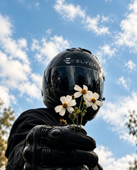 A person in a black 'ELYON' motorcycle helmet and gloves holds a small bouquet of white flowers against a blue, cloudy sky.