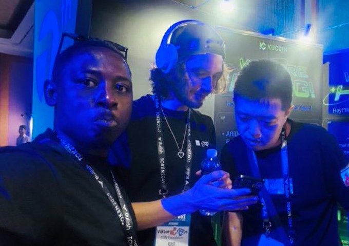 Two people at what appears to be a TOKEN2049 conference (visible on their lanyards) gather around a phone screen, with a "KUCOIN" banner in the background, capturing a casual birthday moment for @0xJeribond.
