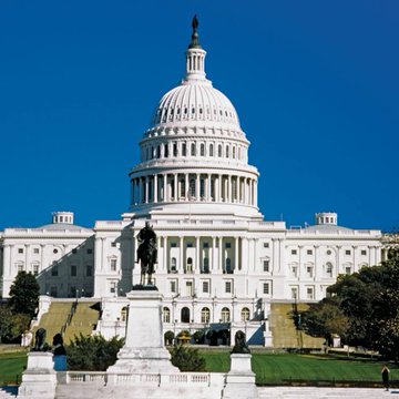 The U.S. Capitol Building stands prominently under a clear blue sky, with a statue visible in the foreground on the Capitol grounds. The image directly relates to the post about the CLARITY Act, as the Capitol is where the U.S. Senate must act to pass the legislation before their summer recess.