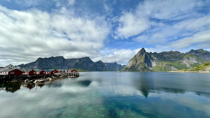 A small cluster of red rorbu cabins sits along the shore of a calm fjord, with dramatic mountain peaks rising under a partly cloudy sky — likely a scene from Norway's Lofoten Islands. The image perfectly captures the essence of "touching grass succeeded too well," offering a serene escape that makes the prospect of returning to work feel even more reluctant.