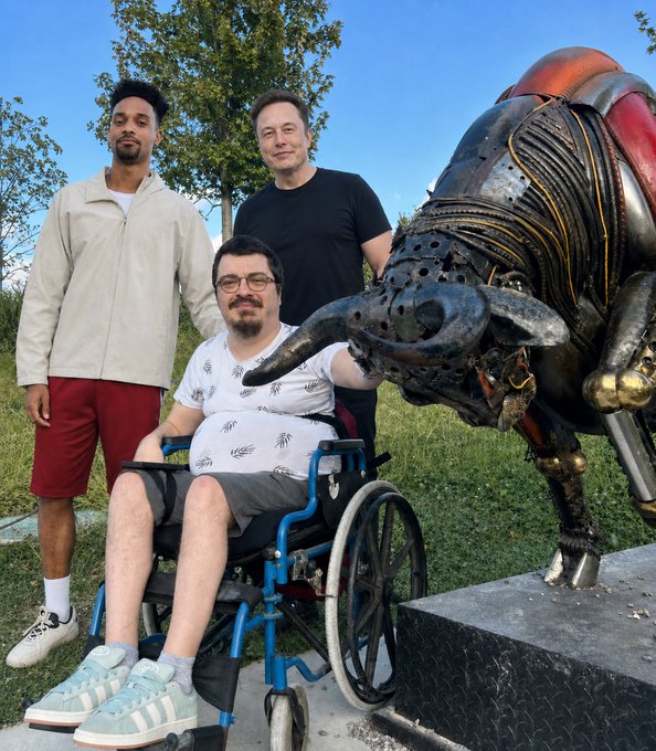 The image shows a young man in a wheelchair posing alongside Elon Musk and another individual next to a large metallic bull sculpture, illustrating his tweet about hoping to undergo Neuralink surgery. The post expresses his dream of receiving the brain-chip implant procedure from Musk's company to improve his quality of life before his parents age.
