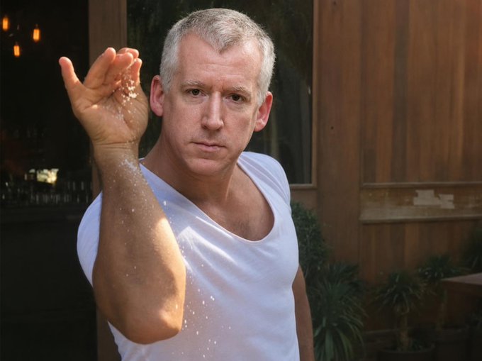 A man with short gray hair wearing a white t-shirt stands outdoors, sprinkling white powder (likely salt or sugar) from his raised hand, with a serious expression on his face.