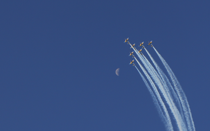Seven airplanes fly in formation leaving white smoke trails against a clear blue sky, with a crescent moon visible to the left of the formation.