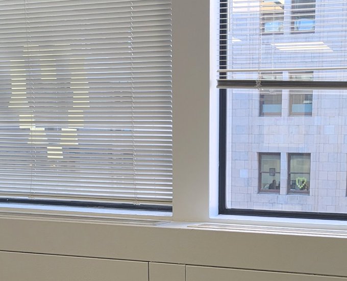 The image shows a window with blinds, allowing light to enter the room. The blinds are white and have a pattern of squares on them. In front of the window, there is a white cabinet or shelf that appears to be made of wood. The background features a building, which could be an office space or another type of commercial establishment.