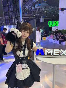 A young woman in a black and white maid outfit poses at the MEX Global Exchange (MEXC) booth, holding up a small plush cat while making a cute gesture. Visible signage includes "MEXC" prominently displayed on the booth and "LOUR" in the background, tying the image to the accompanying post's playful "say meow" theme.