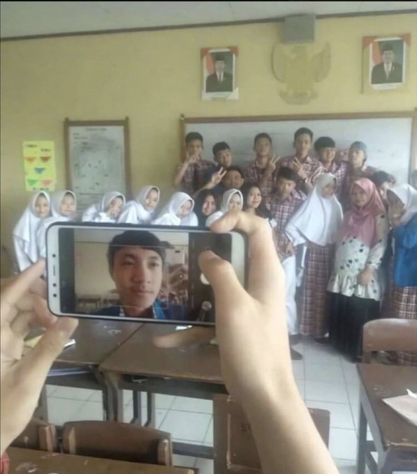 Photo taken on a Samsung smartphone of a classroom group selfie, with Indonesian and presidential flag emblems visible on the wall behind students wearing hijabs and school uniforms.