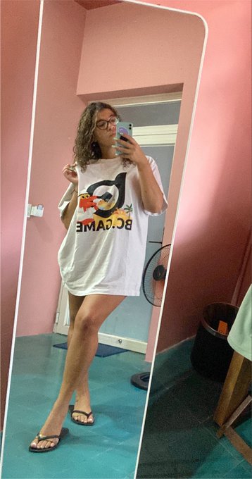 A woman takes a mirror selfie while wearing a white oversized t-shirt featuring a mirrored 'BC GAME' logo, standing in a room with pink walls.