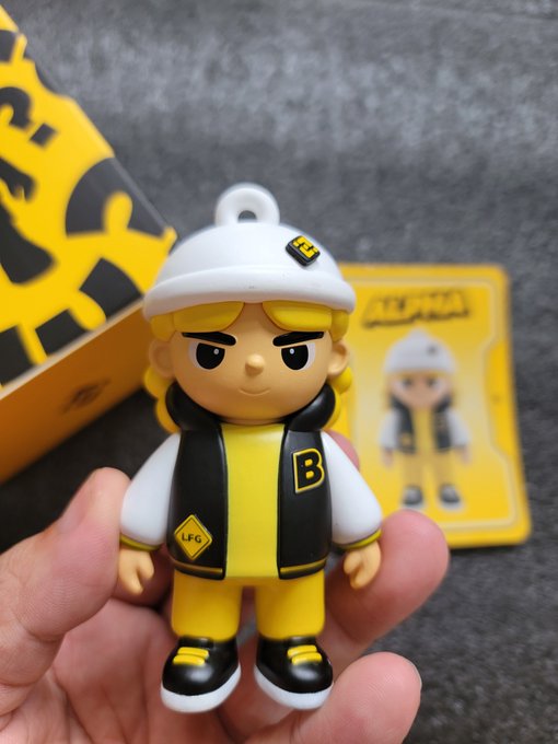 A small collectible figurine of a blonde character wearing a white beanie, black and yellow jacket with 'B' logo, and yellow pants, branded with 'LFG' patches, held in a hand with packaging visible in the background.