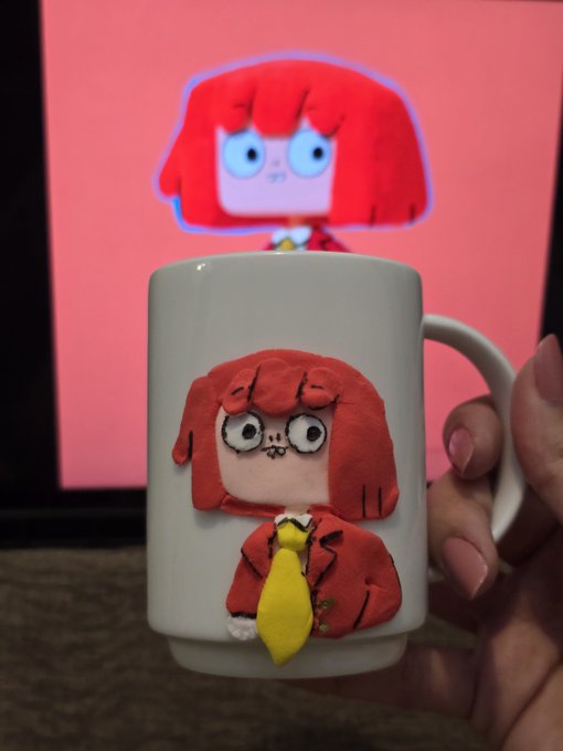 A handmade clay figurine of a red-haired anime-style character sits on a white ceramic mug being held by a person, with the character's animated counterpart displayed on a screen in the background.