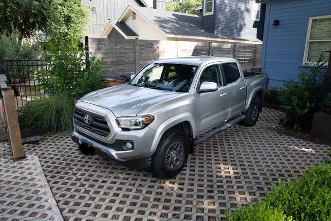 A silver Toyota Tacoma pickup truck is parked on a permeable brick driveway in front of a house with blue siding and a wooden fence.