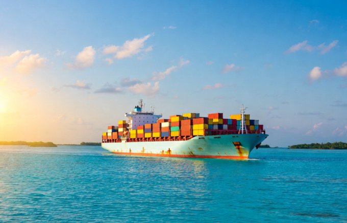 A large cargo container ship loaded with multicolored containers sails on turquoise water during a vibrant sunset.