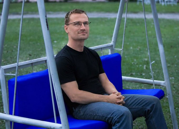 A man wearing glasses and a black t-shirt sitting on a blue cushioned swing in a grassy area.