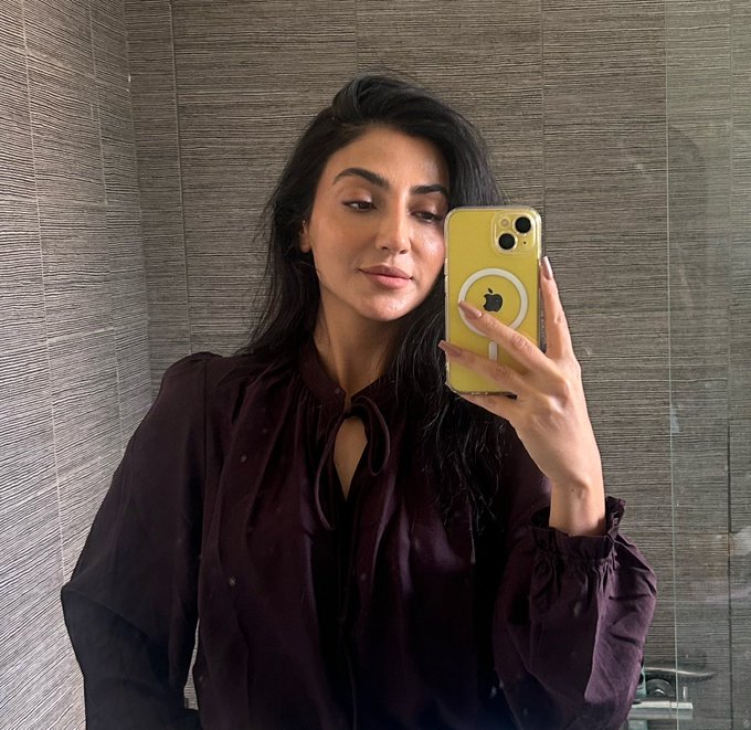 A woman with long dark hair takes a mirror selfie in what appears to be a bathroom with textured tile walls, holding a yellow iPhone with an Apple logo on the case. She pairs the casual "Gm frenz" morning greeting with her relaxed, fresh-faced look, embodying a simple and natural "good morning" vibe.