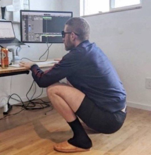 A man squats barefoot at a desk in front of a computer monitor displaying what appears to be audio editing software, with no chair present—visually illustrating the caption "Sold my chair to buy the dip!" by jokingly suggesting he sold his office chair to invest in a cryptocurrency market downturn.