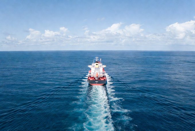 Aerial photograph of a red and white cargo ship sailing across a calm blue ocean under a partly cloudy sky, leaving a foamy wake behind it.