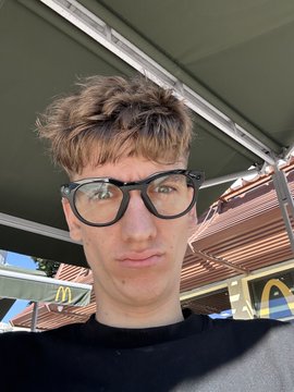 A selfie of a young man with messy brown hair and black-rimmed glasses, wearing a dark sweater, taken in front of a McDonald's sign.