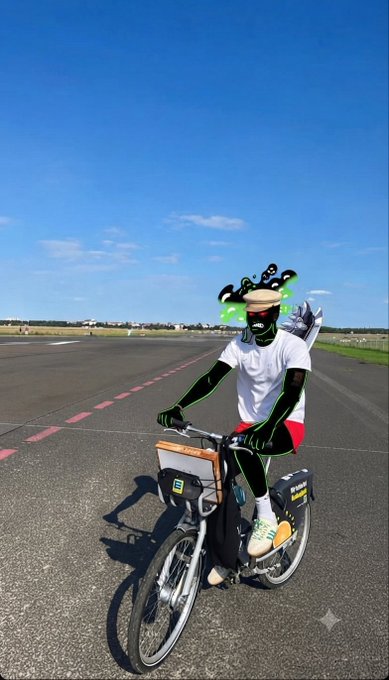 A cyclist on what appears to be a runway or tarmac wearing a white t-shirt, red shorts, green gloves, and a green-tinted mask with a whimsical face graphic.