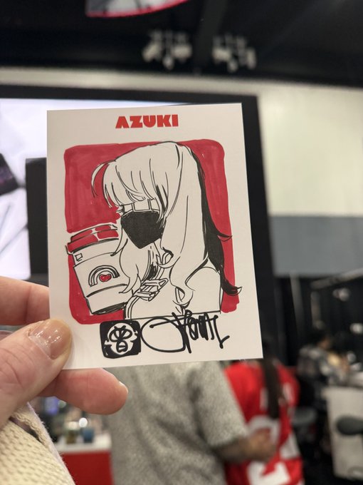 A hand holds an autographed card featuring an anime-style character named 'AZUKI' wearing a face mask, with a coffee cup logo and signature.