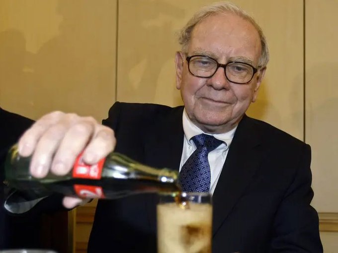 An elderly man with glasses wearing a dark suit and tie, smiling slightly while pouring Coca-Cola from a glass bottle into a glass, with a Coca-Cola label visible on the bottle.
